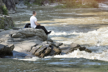 Young man sitting on the bank of a stormy river. Sunny summer day