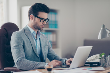 Portrait of hardworking director in shirt and jacket with modern hairstyle searching information on computer, using electronic device, sitting in work place at his desk