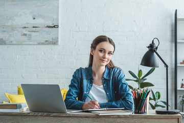 teenage girl taking notes and smiling at camera while studying with laptop at home