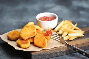 Chicken nuggets and French fries on a wooden board. Next cup with ketchup. One of the nuggets is pinned on the fork. Close-up. Gray background.