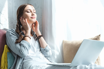 beautiful teen girl using laptop and listening music with earphones on windowsill