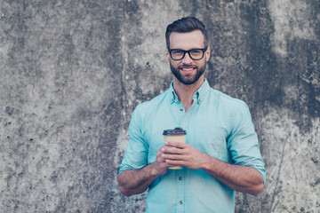 Portrait with copy space of cheerful attractive man with modern hairstyle holding carton cup with coffee in hands looking at camera isolated on grey stone background. Advertisement concept