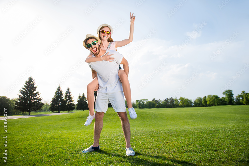 Poster Full size fullbody portrait of cheerful positive couple man carrying on back redhead lover showing two fingers v-sign over green grass. Field blue sky sunlight weekend vacation holidays concept