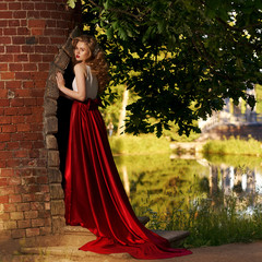 Fototapeta premium Fashion style photo of young beautiful woman in evening dress with white top and red flying skirt posing in summer park at sunset under green trees and close to old brick tower.