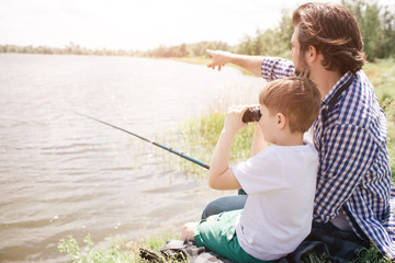 Boy is sitting with his dad at the river shore and looking through binoculars. Adult man is pointing forward and holding fish-rod in right hand.