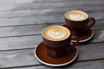Two dark red cups of cappuccino with the heart shape latte-art on wood background