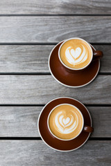 Two dark red cups of cappuccino with the heart shape latte-art on wood background