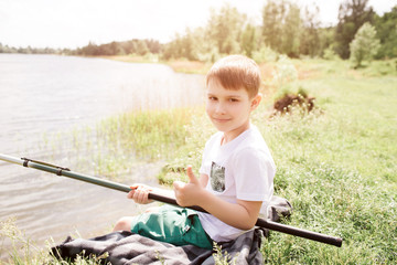 Small boy is sitting on blanket at river shore alone. He is holding fish-rod with both hands and looking at camera. He is enjoying the moment.