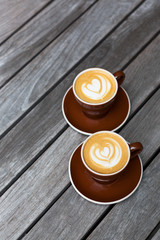 Two dark red cups of cappuccino with the heart shape latte-art on wood background