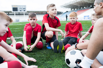 Portrait of group of eager teenage boys sitting in circle listening to motivational pep talk from coach before team practice in outdoor stadium