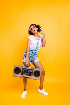 Full body portrait of positive laughing girl in eyewear jumpsuit holding boom box in hand gesturing rock and roll sign isolated on yellow background. Party maker music lover fan hobby concept