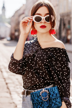 Outdoor portrait of young beautiful fashionable woman wearing stylish white oval sunglasses, red tassel earrings, polka dot blouse, blue waist bag, posing in street of city. Summer fashion concept