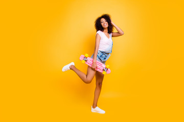 Fototapeta na wymiar Portrait of cute cheerful coquette holding pink skate board in hand posing with raised leg enjoying weekend holiday isolated on yellow background