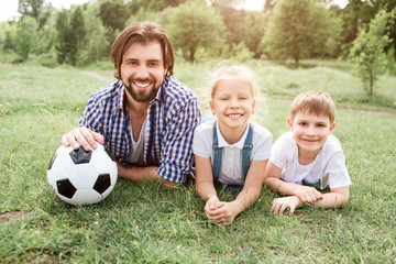A picture of father lying on grass with his kids. He is holding ball besides himself. They are looking on camera amd smiling.