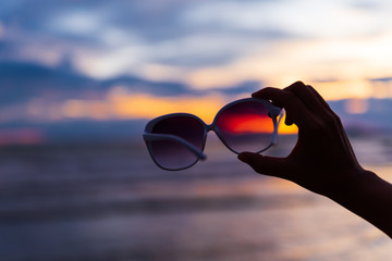 Silhouette woman hand holding sunglasses over sea and Sandy beach in background during sunset for...