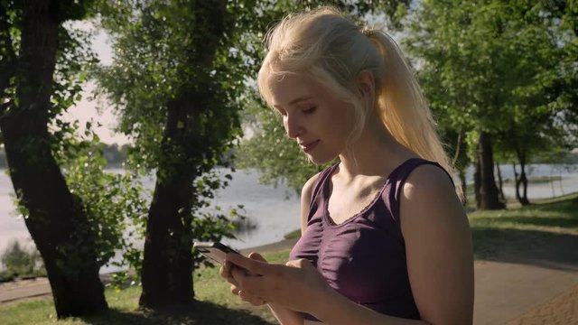 Young blonde woman with ponytail typing on her phone and looking in camera, smiling, park near river background