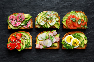 Avocado sandwiches, toasts with various vegetarian toppings on a black stone background, top view