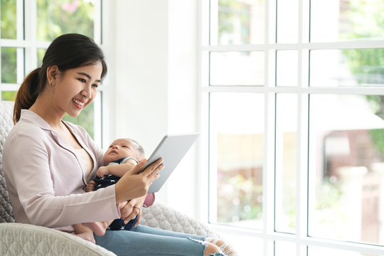 Happy attractive asian mother with kid sitting at couch in living room searching parents education in social network or internet via smart tablet. Young parents lifestyle with copy space concept. - Powered by Adobe