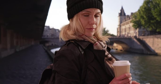 Casual blonde woman traveling in Paris looking gently at camera by Seine River, Attractive female tourist in Paris, France in coat and beanie staring directly at camera, 4k