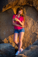 Beautiful young girl standing, leaning against a stone wall at sunset with a cocktail in her hand