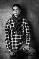 Portrait of young trendy handsome man with short dark hair wearing checkered shirt and brown trousers standing and posing against gray concrete wall