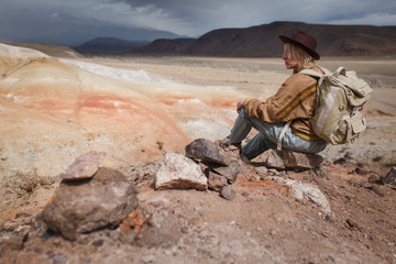 Beautiful young girl in hare, hat and with backpack siting among sands in wild