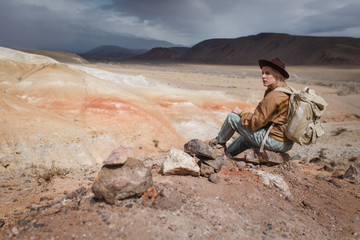 Beautiful young girl in hare, hat and with backpack siting among sands in wild