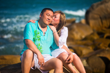 Adult Asian woman kissing her man sitting on the rocks, on the sea background
