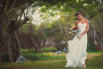 Beautiful young bride posing in a wedding dress in the middle of tropical trees and looks at the wedding bouquet