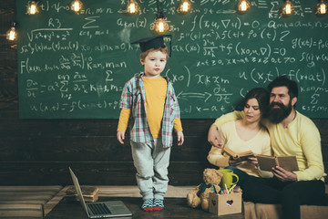 Smart child in graduate cap like to perform. Parents listening their son, checking mistakes with book chalkboard on background. Preparing for exam concept. Boy presenting his knowledge to mom and dad