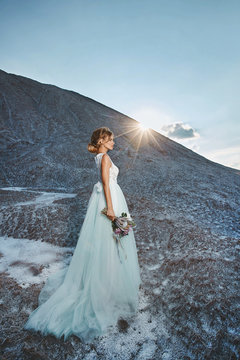 Fashionable and beautiful blonde model girl with modeling hairstyle in stylish white lace dress with bouquet of exotic flowers in her hand posing on the mountain at the sunset