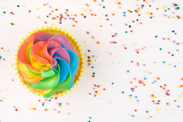 Cupcake with rainbow colorful cream in yellow cup on white wooden table decorated with colorful sprinkles. Top view.