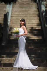 Naklejka premium A young bride in a beautiful white dress stands with a bouquet of flowers on the stone steps