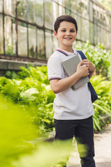 smiling schoolboy with backpack and book looking at camera in garden