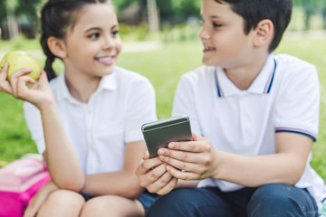 schoolchildren using smartphone together and chatting while sitting on grass