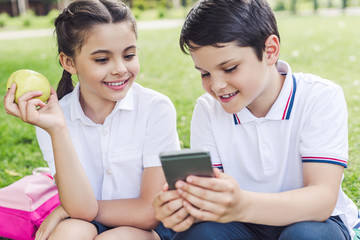 smiling schoolchildren using smartphone together while sitting on grass