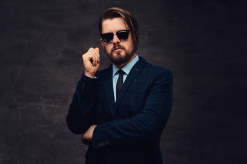 Obraz premium Close-up portrait of a handsome fashionable middle-aged man with beard and hairstyle dressed in an elegant formal suit and sunglasses. Isolated on a textured dark background in studio.