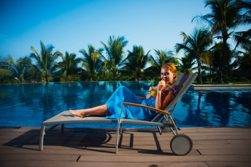 A young girl in a blue dress lying on a sun lounger by the pool and smiling, holding juice