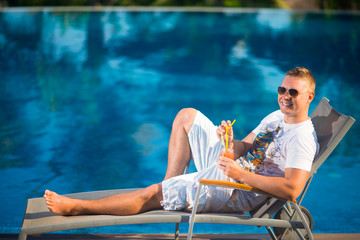 A man in a white t-shirt and sunglasses lying on a lounger by the pool, holding juice