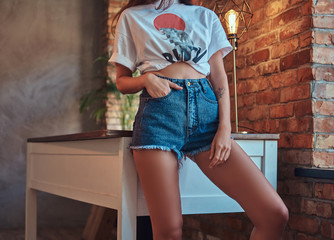 Cropped image of a sexy slim cool girl in a white top and denim shorts, leaning on a table in...