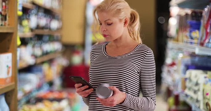 Woman at the grocery store checking nutrition facts label on can of tuna, Portrait of mom with her phone shopping at the supermarket, 4k