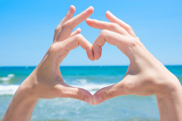 Young Caucasian Woman Girl Making Heart with Hands Palms. Turquoise Sea with Waves Blue Sky Horizon Background. Bright Sunlight. Summer Vacation Travel Wanderlust Love Concept. Poster with Copy Space