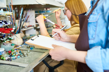 Close up of young woman holding clipboard while colleague in background working with glass in modern glassworking studio, copy space