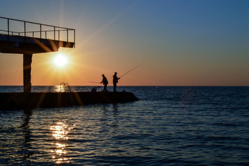 two men stand on the pier and fish with a fishing rod. pier has two tiers. men are fishing against the setting sun