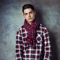 Portrait of young trendy handsome man with short dark hair wearing checkered shirt and brown trousers standing and posing against gray concrete wall