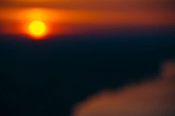 Blurry background image of circle of sun rising from behind dark horizon on background varicolored clouds of warmly shades above river. Abstract picturesque image of sun in bokeh.