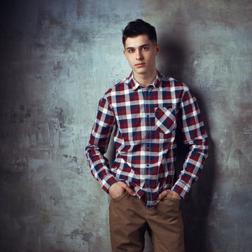 Portrait of young trendy handsome man with short dark hair wearing checkered shirt and brown trousers standing and posing against gray concrete wall