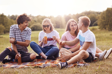 Fototapeta premium Four friendly youngsters spend free time outdoor, sit on plaid, have pleasant talk, drink cider, have pleasant smiles. Two coulpes have picnic outside. Friendship, companionship, relationship concept