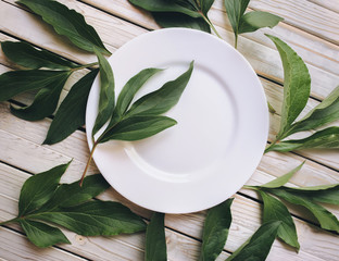 Top view empty white plate on rustic wooden background with green leaves of peony.