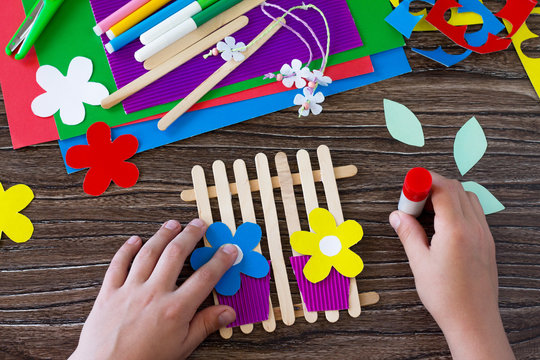 The child glues the details of the gift of summer flowers on wooden sticks fence. Handmade. Project of children's creativity, handicrafts, crafts for kids.